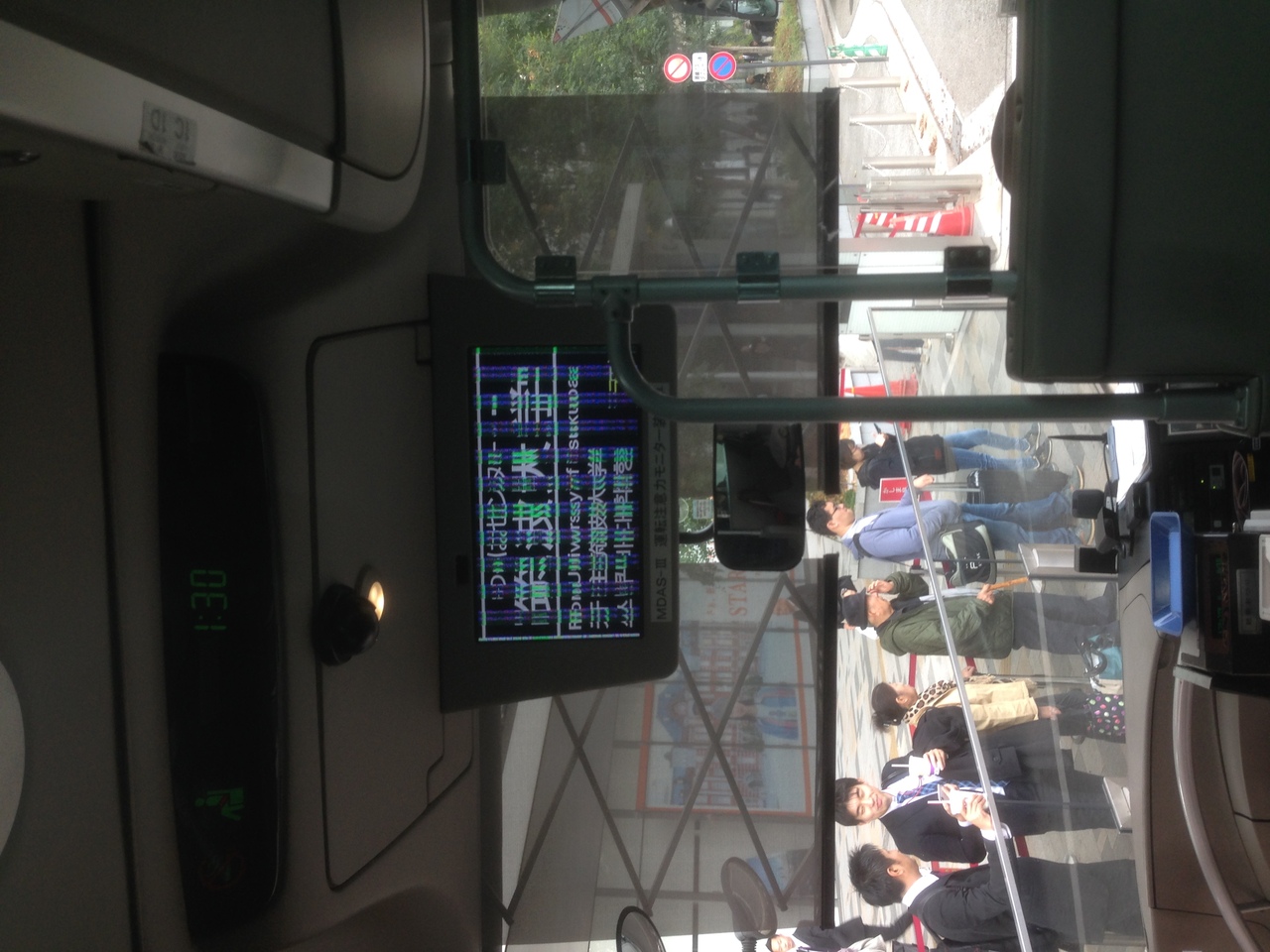 bus monitor showing glitched text in Japanese, people are waiting in a queue outside